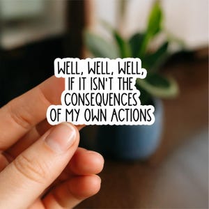 May include: A white sticker with black text saying "WELL, WELL, WELL, IF IT ISN'T THE CONSEQUENCES OF MY OWN ACTIONS." The sticker is held by a hand, with a blurred background of a plant and a dark blue pot.
