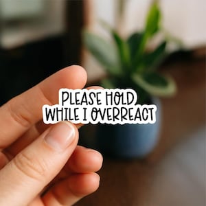 May include: A white sticker with black text that reads "PLEASE HOLD WHILE I OVERREACT". The sticker is held between fingers, with a blurred background of a plant in a dark blue pot.