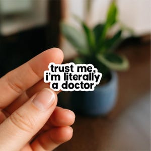 May include: A white sticker with black text that reads "trust me, I'm literally a doctor." The sticker is held in a person's hand, with a blurred background of a plant in a pot.