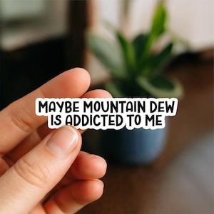 May include: A white sticker with black text that reads "MAYBE MOUNTAIN DEW IS ADDICTED TO ME." The sticker is held by a hand in front of a blurred background with a plant.