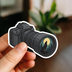May include: A black camera sticker with a lens displaying a wolf howling at the moon in a forest. The sticker is held by a hand and has a white border. The camera is detailed with buttons and a flash.