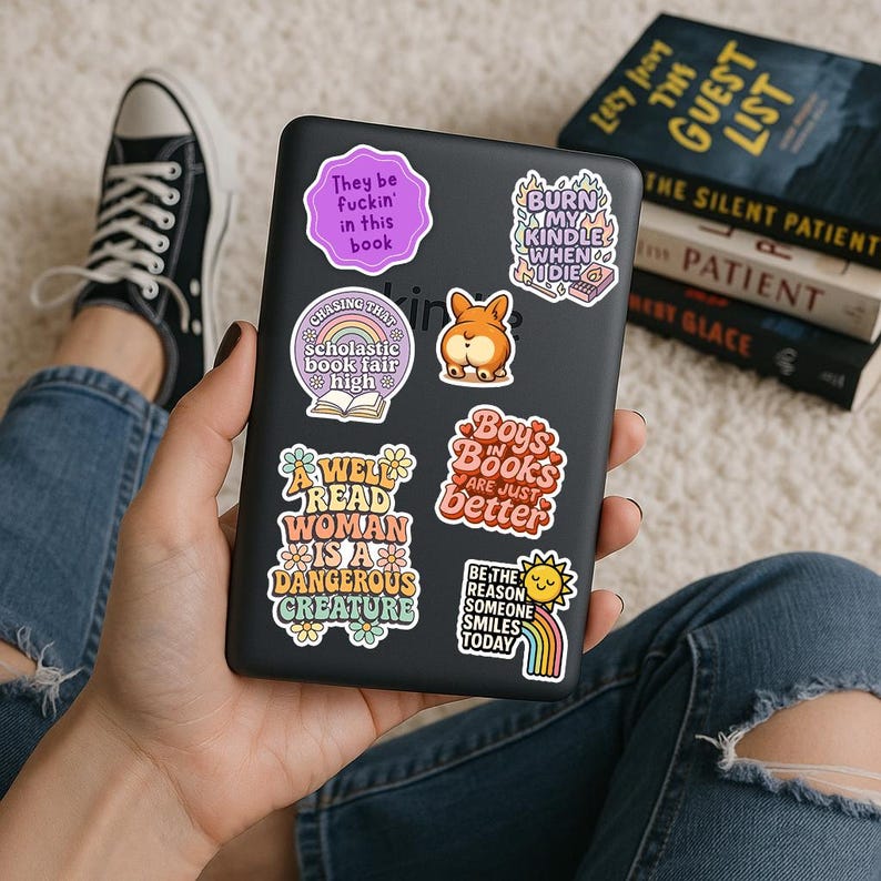 A person is holding a tablet device with various stickers on it, surrounded by books and a pair of sneakers.