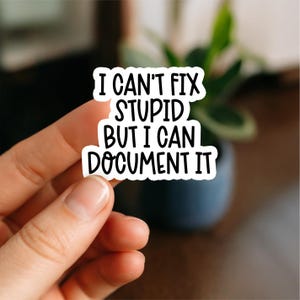 May include: A white sticker with black text that reads "I CAN'T FIX STUPID BUT I CAN DOCUMENT IT." The sticker is held between fingers against a blurred background of a plant and a dark pot.