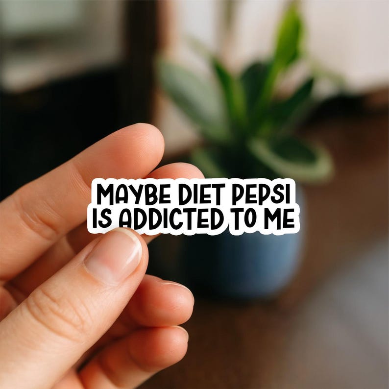 Op de afbeelding: Een witte sticker met zwarte tekst die luidt: "MAYBE DIET PEPSI IS ADDICTED TO ME." De sticker wordt tussen twee vingers gehouden. De achtergrond is wazig, met een plant in een donkerblauwe pot.