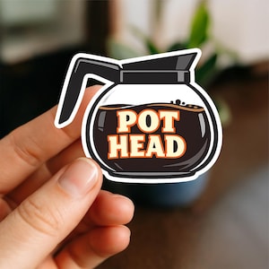 May include: A die-cut sticker shaped like a glass coffee pot with a black handle and lid. The pot is filled with dark brown coffee and features the text POT HEAD in bold, cream-coloured letters with a thin orange outline centred on the front.