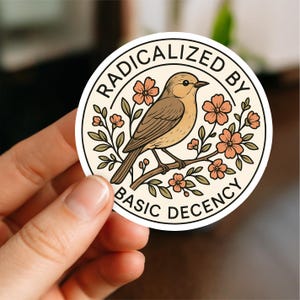 May include: A round sticker with the text "RADICALIZED BY BASIC DECENCY" encircling a bird perched on a flowering branch. The bird is brown and tan, the flowers are peach, and the background is cream.