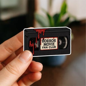 May include: A black VHS tape sticker with the text "HORROR MOVIE FAN CLUB" on a tan label. Red blood drips from the top of the tape. The sticker is held by a person's hand.