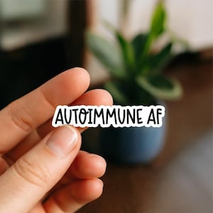 May include: A white sticker with the words "AUTOIMMUNE AF" in black, held between fingers. The background is blurred, featuring a green plant in a dark blue pot and a wooden surface.