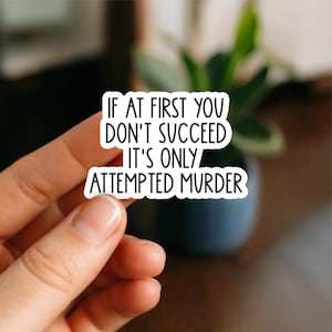 May include: A white sticker with black text that reads "IF AT FIRST YOU DON'T SUCCEED IT'S ONLY ATTEMPTED MURDER." The sticker is held by a person's fingers, with a blurred background of a plant and a dark blue pot.