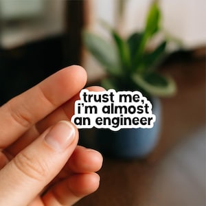 May include: A white sticker with black text that reads "trust me, I'm almost an engineer." The sticker is held between fingers against a blurred background. The text is in a sans-serif font.