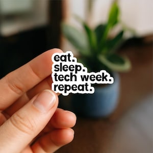 May include: A white sticker with black text that reads "eat. sleep. tech week. repeat." The sticker is held by a hand, with a blurred background of a plant in a dark blue pot.