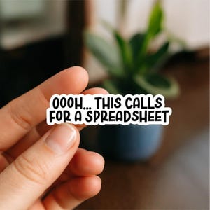 May include: A white sticker with black text that reads "OOOH... THIS CALLS FOR A SPREADSHEET". The sticker is held by a hand, with a blurred background of a plant and a dark surface.