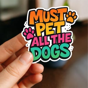 May include: A vibrant sticker with the phrase "MUST PET ALL THE DOGS" in a fun font. The design includes paw prints and a gradient of orange, yellow, pink, and green. The sticker is held in a hand.