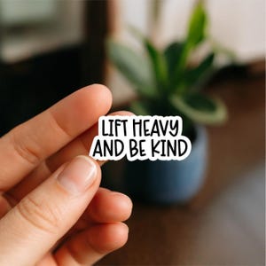 May include: A white sticker with black text that reads "LIFT HEAVY AND BE KIND." The sticker is held by a hand, with a blurred background featuring a plant in a dark blue pot. The sticker is likely for a water bottle, laptop, or other personal items.
