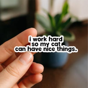 May include: A white sticker with black text that reads "i work hard so my cat can have nice things." The sticker is held between fingers, with a blurred background of a plant and a dark blue pot.