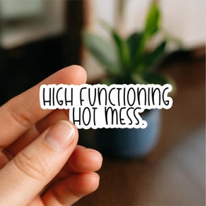 Könnte beinhalten: Ein weißer Aufkleber mit dem schwarzen Text "HIGH FUNCTIONING HOT MESS". Der Aufkleber wird von einer Hand gehalten, mit einem verschwommenen Hintergrund, der eine Pflanze in einem dunklen Topf zeigt.