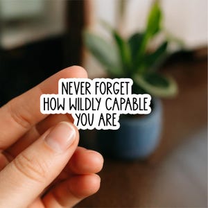 May include: A white sticker with black text that reads "NEVER FORGET HOW WILDLY CAPABLE YOU ARE." The sticker is held in front of a blurred background with a plant and a dark blue pot.