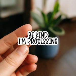 May include: A white sticker with black text that reads "BE KIND I'M PROCESSING." The sticker is held between fingers, with a blurred background of a plant in a dark blue pot.
