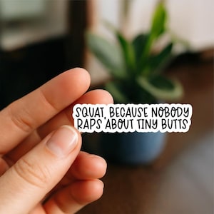 May include: A white sticker with black text that reads "SQUAT, BECAUSE NOBODY RAPS ABOUT TINY BUTTS." The sticker is held between fingers in front of a blurred background with a plant.