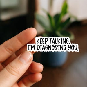May include: A white sticker with black text that reads "KEEP TALKING. I'M DIAGNOSING YOU." The sticker is held in a person's hand, with a blurred background of a plant and a dark blue pot.