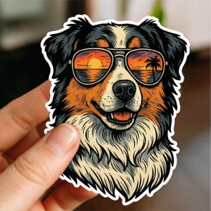 Peut inclure: Un autocollant représentant un chien portant des lunettes de soleil reflétant un coucher de soleil sur l'océan avec un palmier. Le chien a des poils noirs, bruns et blancs. L'autocollant est tenu par une main.