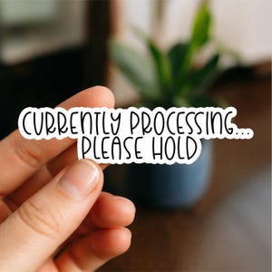 May include: A white sticker with black text saying "CURRENTLY PROCESSING... PLEASE HOLD". The sticker is held by a hand, with a blurred background of a plant and a dark blue pot.