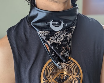 May include: A black sleeveless shirt with a gold circular design featuring an eye and a pendant necklace with a white stone. The shirt has a black and white paisley bandana with a black crescent moon and a white star design around it.