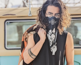 May include: A person wearing a black sleeveless shirt with a black bandana featuring a white moon, eye, and mushrooms. The bandana has the text "Shroom Queens" on it. The person is wearing glasses and has long, curly hair.
