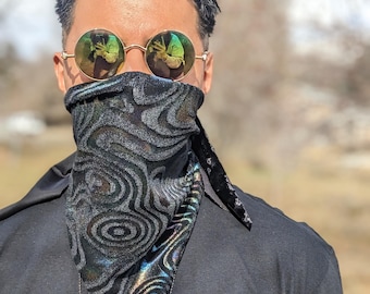 May include: A person wearing a black t-shirt with a silver chain necklace and a black bandana with a swirling iridescent pattern. The bandana covers the person's mouth and nose. The person is wearing round, gold-rimmed sunglasses with green mirrored lenses.