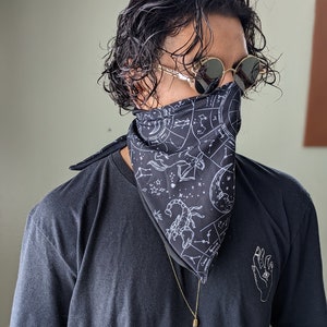 May include: A person wearing a black t-shirt with a white constellation print bandana and a gold necklace with a crystal pendant. The person is also wearing round, gold-rimmed sunglasses.