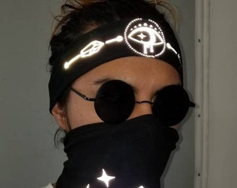 May include: A black bandana with a reflective white diamond design and two stars. The bandana is worn by a person with a black headband with a reflective white eye design.
