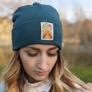 May include: A teal blue beanie with a colorful sunset graphic patch. The patch features a sun, clouds, and a landscape with wavy lines.