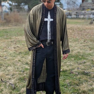 May include: A long, olive green robe with black trim and a black t-shirt with a white graphic of a sword. The robe has a subtle striped pattern and is worn over black jeans and brown boots. The outfit is worn outdoors.