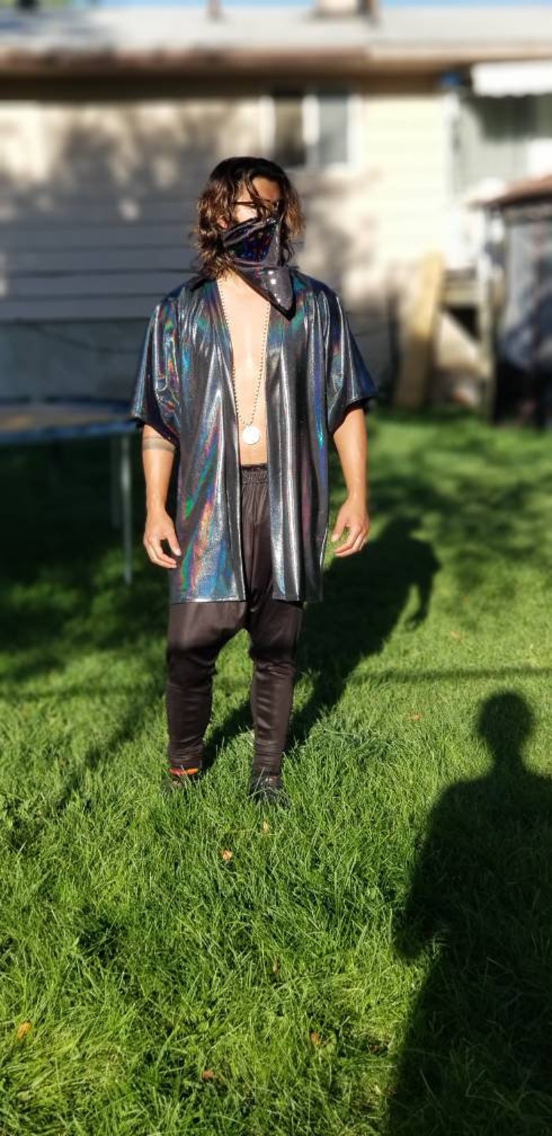 May include: A person wearing a black iridescent shirt with a black bandana over their face. The shirt has a silver chain necklace with a round pendant. The person is standing in a grassy area.