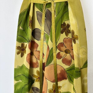 K&ouml;nnte beinhalten: Ein flie&szlig;endes, gelbes Blumenkleid mit Bindedetail in der Taille. Das Kleid hat ein lebendiges Muster aus gro&szlig;en orangefarbenen und braunen Blumen, gr&uuml;nen Bl&auml;ttern und kleineren gelben Bl&uuml;ten. Das Kleid hat einen weichen, flie&szlig;enden Stoff und eine passende gelbe Schleife.