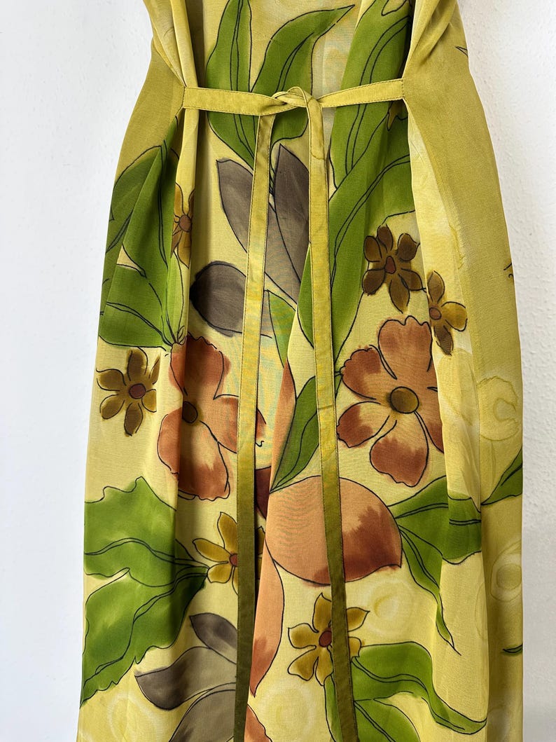 K&ouml;nnte beinhalten: Ein flie&szlig;endes, gelbes Blumenkleid mit Bindedetail in der Taille. Das Kleid hat ein lebendiges Muster aus gro&szlig;en orangefarbenen und braunen Blumen, gr&uuml;nen Bl&auml;ttern und kleineren gelben Bl&uuml;ten. Das Kleid hat einen weichen, flie&szlig;enden Stoff und eine passende gelbe Schleife.