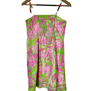 Op de afbeelding: Een roze zomerjurk met een groen en roze olifanten- en palmbomenpatroon. De jurk heeft een gesmokte lijfje en spaghettibandjes. De jurk hangt aan een houten kleerhanger.