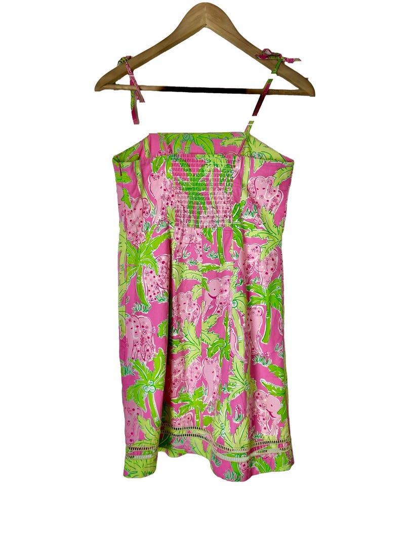 Op de afbeelding: Een roze zomerjurk met een groen en roze olifanten- en palmbomenpatroon. De jurk heeft een gesmokte lijfje en spaghettibandjes. De jurk hangt aan een houten kleerhanger.