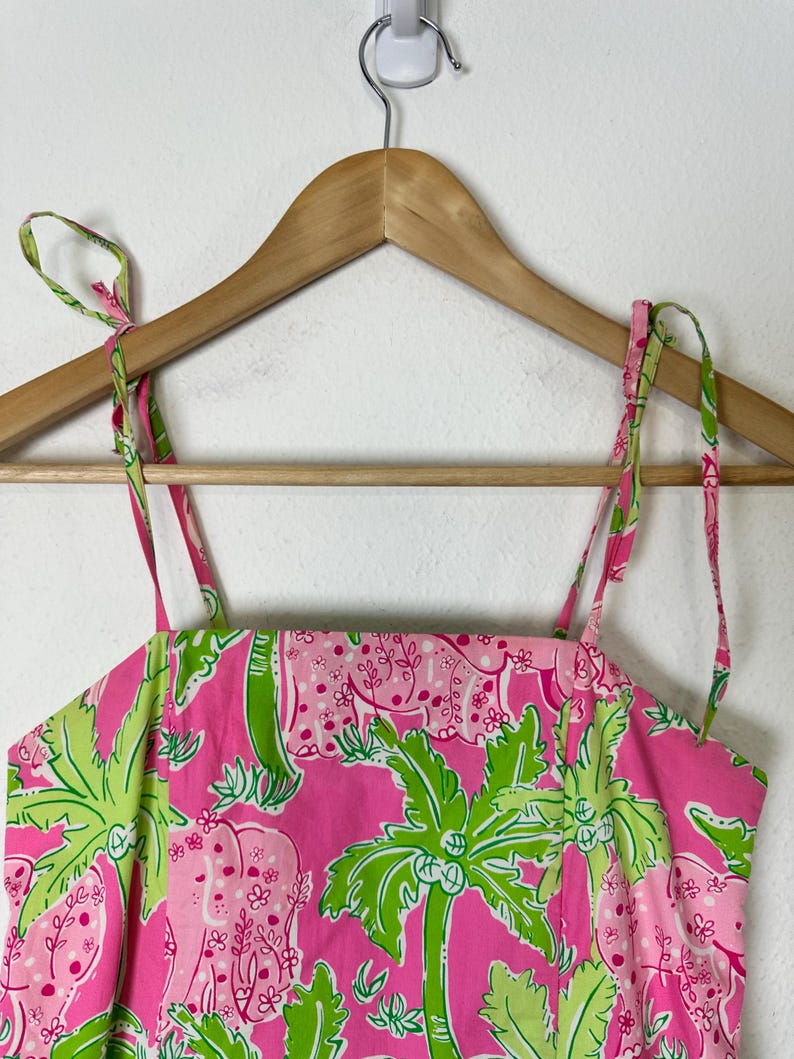 Op de afbeelding: Roze zomerjurk met een tropisch patroon van groene palmbomen en roze olifanten. De jurk heeft dunne, verstelbare bandjes en hangt aan een houten hanger.