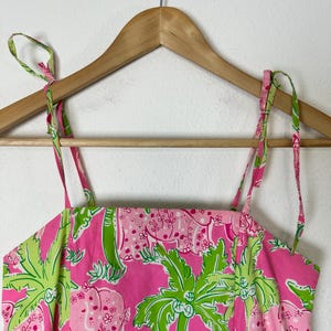 May include: A pink sundress with a tropical print featuring elephants and palm trees. The dress has thin, adjustable straps and is hanging on a wooden hanger. The fabric is a vibrant pink with green and white accents.