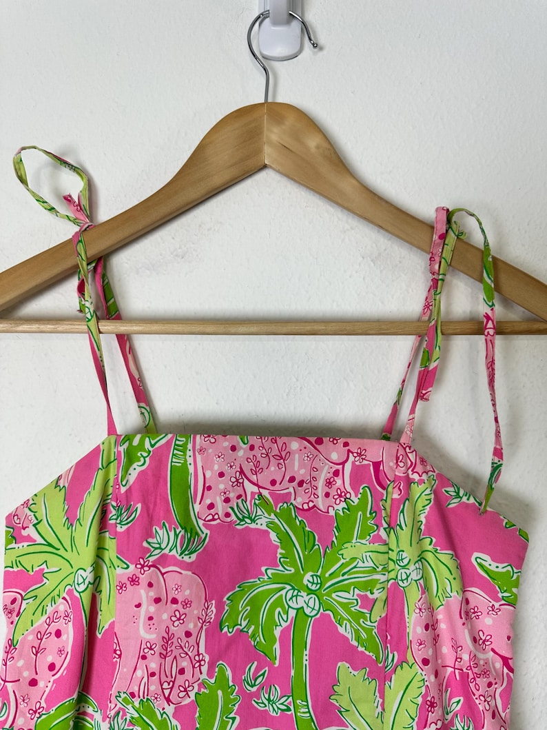May include: A pink sundress with a tropical print featuring elephants and palm trees. The dress has thin, adjustable straps and is hanging on a wooden hanger. The fabric is a vibrant pink with green and white accents.