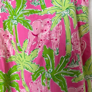 Op de afbeelding: Close-up van een roze stof met een tropisch patroon. Het ontwerp toont roze olifanten, groene palmbomen en kleine witte bloemen tegen een felroze achtergrond. De stof heeft een levendige en speelse uitstraling.