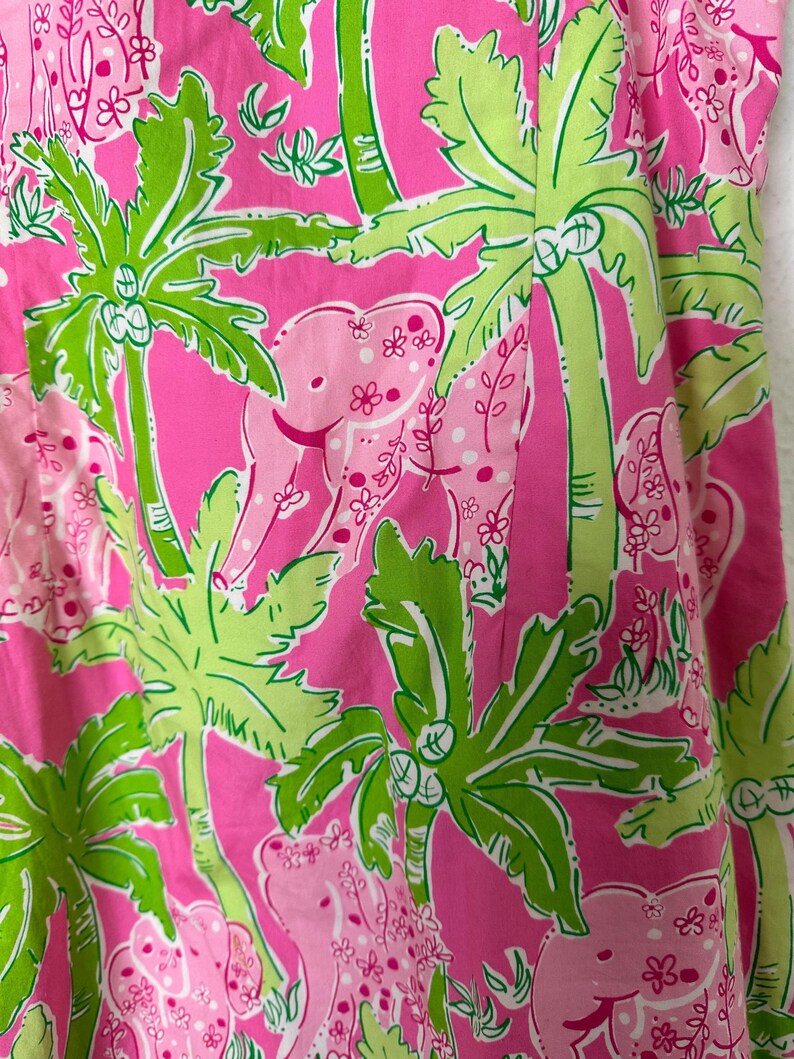 Op de afbeelding: Close-up van een roze stof met een tropisch patroon. Het ontwerp toont roze olifanten, groene palmbomen en kleine witte bloemen tegen een felroze achtergrond. De stof heeft een levendige en speelse uitstraling.