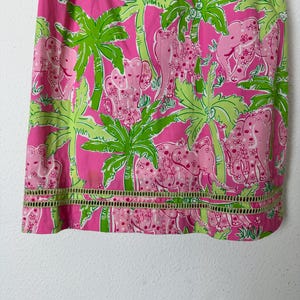 May include: Pink fabric with a repeating pattern of pink elephants and green palm trees. The fabric has a decorative cut-out border at the bottom. The design is bright and colorful, with a tropical theme.