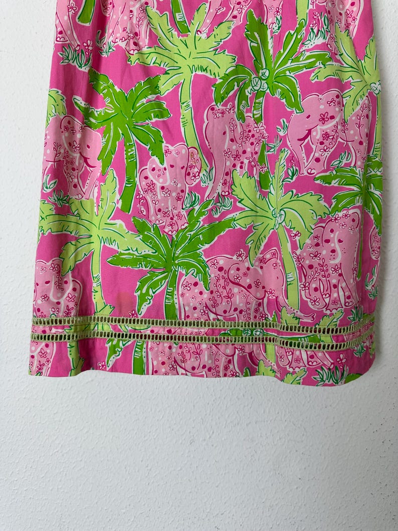 May include: Pink fabric with a repeating pattern of pink elephants and green palm trees. The fabric has a decorative cut-out border at the bottom. The design is bright and colorful, with a tropical theme.