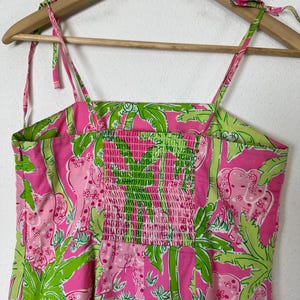 May include: A pink sundress with a green and pink tropical print featuring elephants and palm trees. The dress has adjustable tie straps and a smocked bodice. The dress is hanging on a wooden hanger.