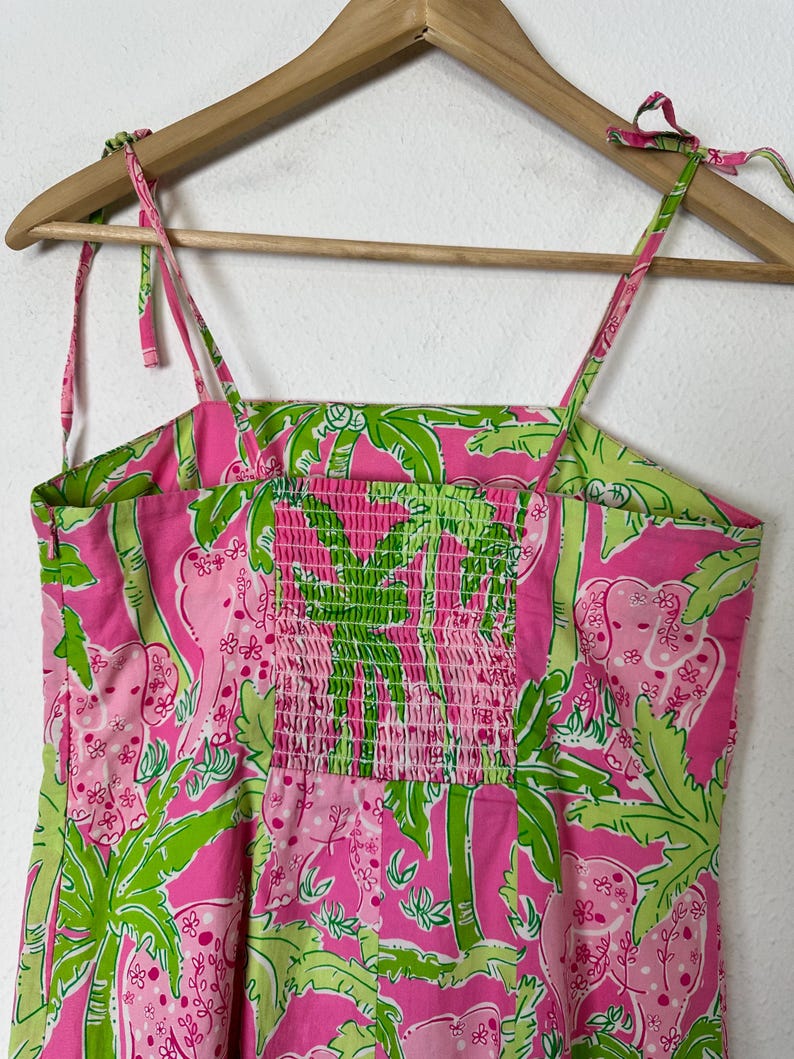 May include: A pink sundress with a green and pink tropical print featuring elephants and palm trees. The dress has adjustable tie straps and a smocked bodice. The dress is hanging on a wooden hanger.