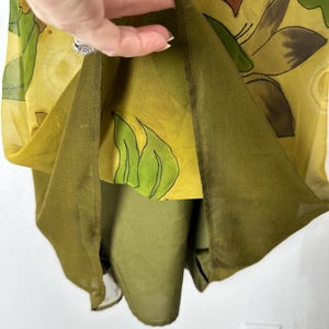 K&ouml;nnte beinhalten: Ein mehrschichtiges Kleid mit gelbem Blumendruck &uuml;ber einer einfarbigen olivgr&uuml;nen Unterschicht. Die &auml;u&szlig;ere Schicht ist transparent, mit braunen und gr&uuml;nen Blumendesigns. Das Kleid scheint aus einem leichten Stoff zu sein.