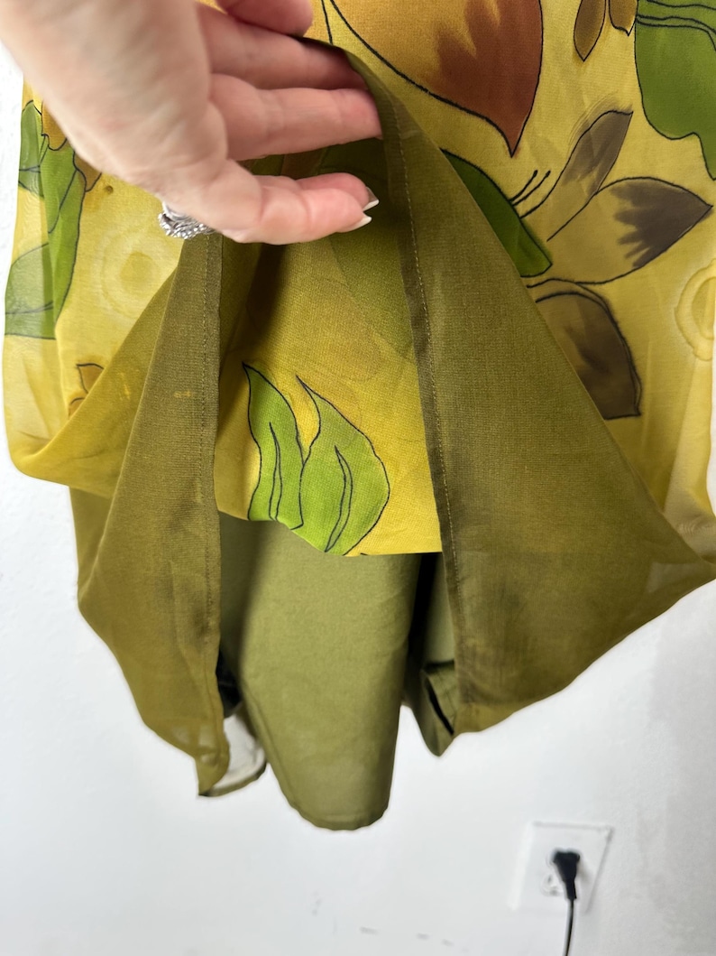 K&ouml;nnte beinhalten: Ein mehrschichtiges Kleid mit gelbem Blumendruck &uuml;ber einer einfarbigen olivgr&uuml;nen Unterschicht. Die &auml;u&szlig;ere Schicht ist transparent, mit braunen und gr&uuml;nen Blumendesigns. Das Kleid scheint aus einem leichten Stoff zu sein.