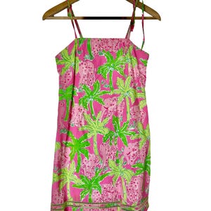 May include: Pink sundress with a tropical pattern of pink elephants and green palm trees. The dress has tie straps and a scalloped hem. The dress is hanging on a wooden hanger.